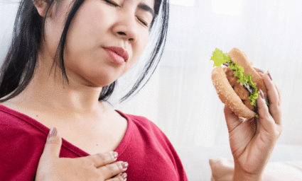 žena s hamburgerem v ruce, držící se za prsa, protože ji trápí reflux.