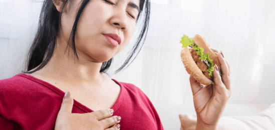 žena s hamburgerem v ruce, držící se za prsa, protože ji trápí reflux.